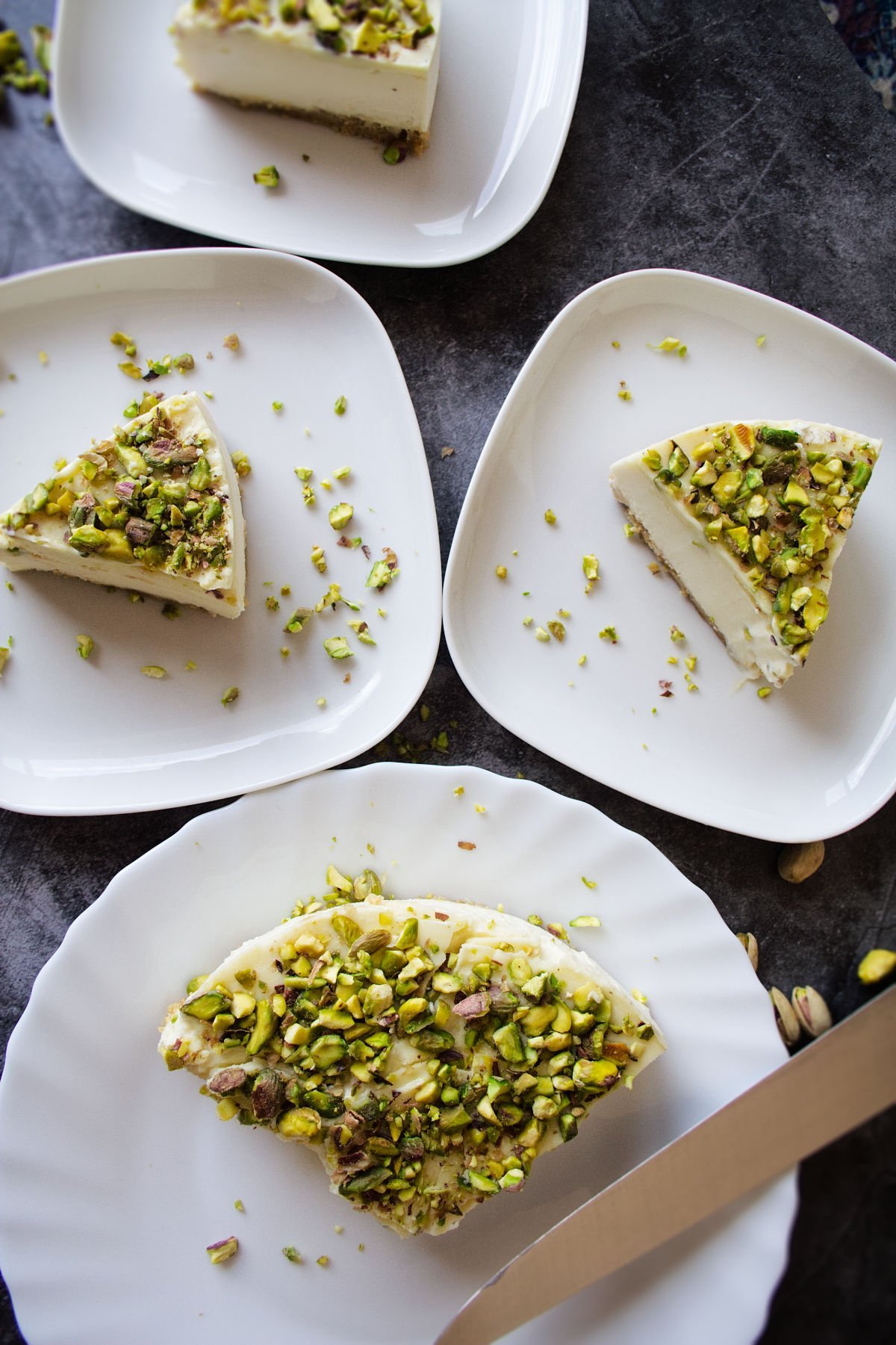 Slices of white chocolate cheesecake with pistachio crust sit on a few. plates waiting to be served.
