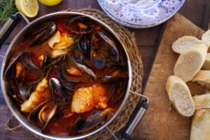 A large pot of Mediterranean fish stew sits beside some sliced bread and some lemons
