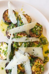 Some Spanish garlic zucchinis sit on a white plate topped with shavings of parmesan cheese