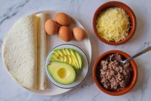 ingredients required for making a tortilla breakfast wrap are laid out on a white granite table