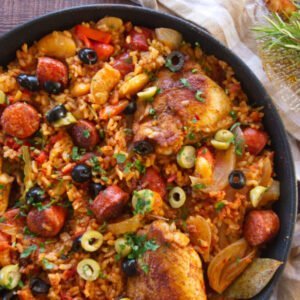 A large pan of Spanish chicken and rice sits garnished with olives and fresh chopped parsley