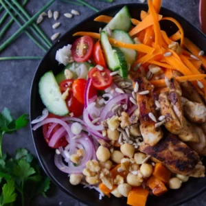 A grilled chicken rice bowl sits on a table with plenty of herbs and sald toppings.