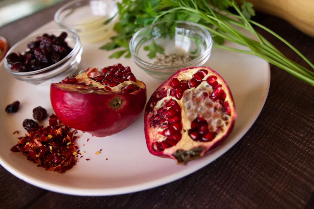 A fresh pomegranate is browkn open on a plate beside some chili flakes, salt, and fresh parsley.