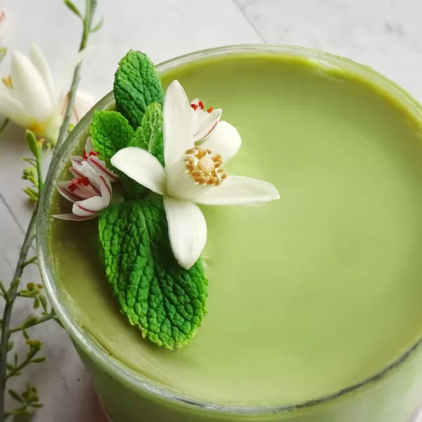 a small glass bowl of avocado cheesecake sits with a few sprigs of fresh mint and an orange blossom.