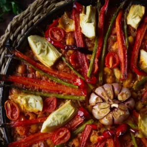a large pan of vegan arroz al horno is topped with roasted red pepper and a whole head of garlic