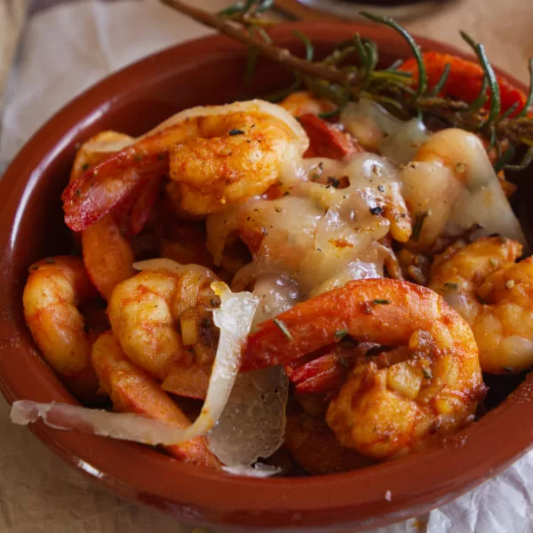 A small earthenware dish full of Spanish garlic shrimp is topped with Manchego cheese and a sprig of rosemary