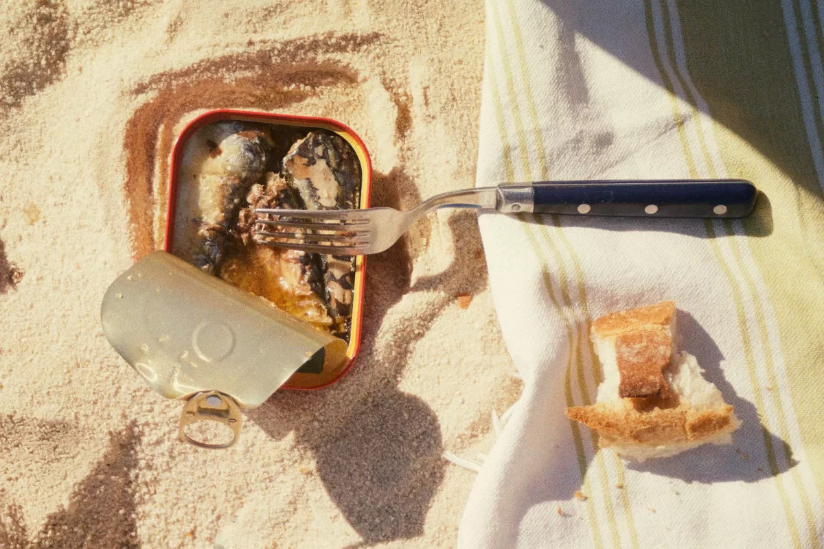 An opened tin of sardines sits on a white sand beach next to a picnic rug. 