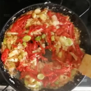 grated tomato and some roasted bell pepper are added to a pan of onion and leek.