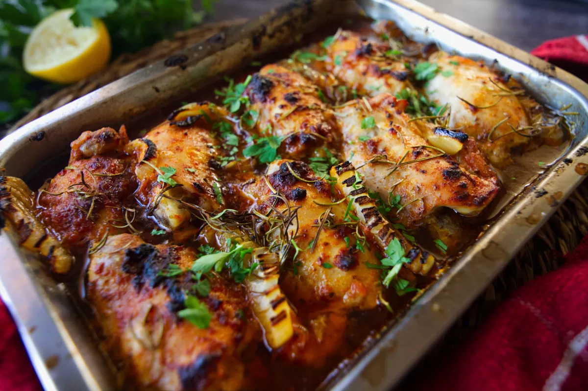 A baking tray of Mediterranean chicken thighs sits beside some fresh parsley and some lemon