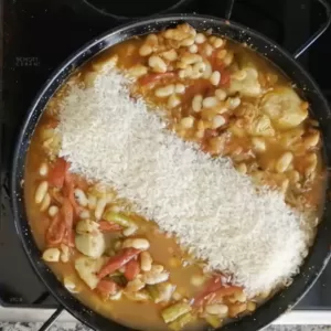 Rice is added to a pan of sauteed vegetables and sofrito sauce