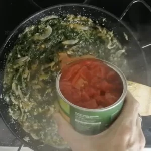 A large can of tomatoes is added to a pan of sauteed spinach