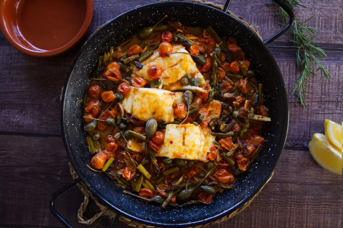 a few fillets of cod fish sit in a pan with sauteed asparagus and cherry tomatoes.