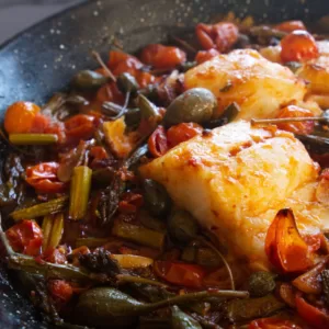 a few fillets of cod fish sit in a pan with sauteed asparagus and cherry tomatoes.