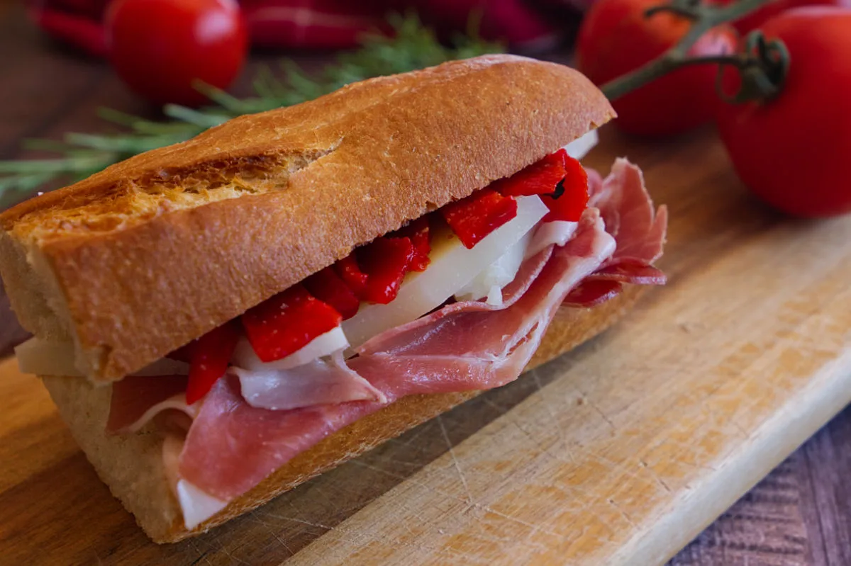 A Jamón Serrano and Manchego cheese bocadillo sits on a wooden chopping board beside some ripe tomatoes.