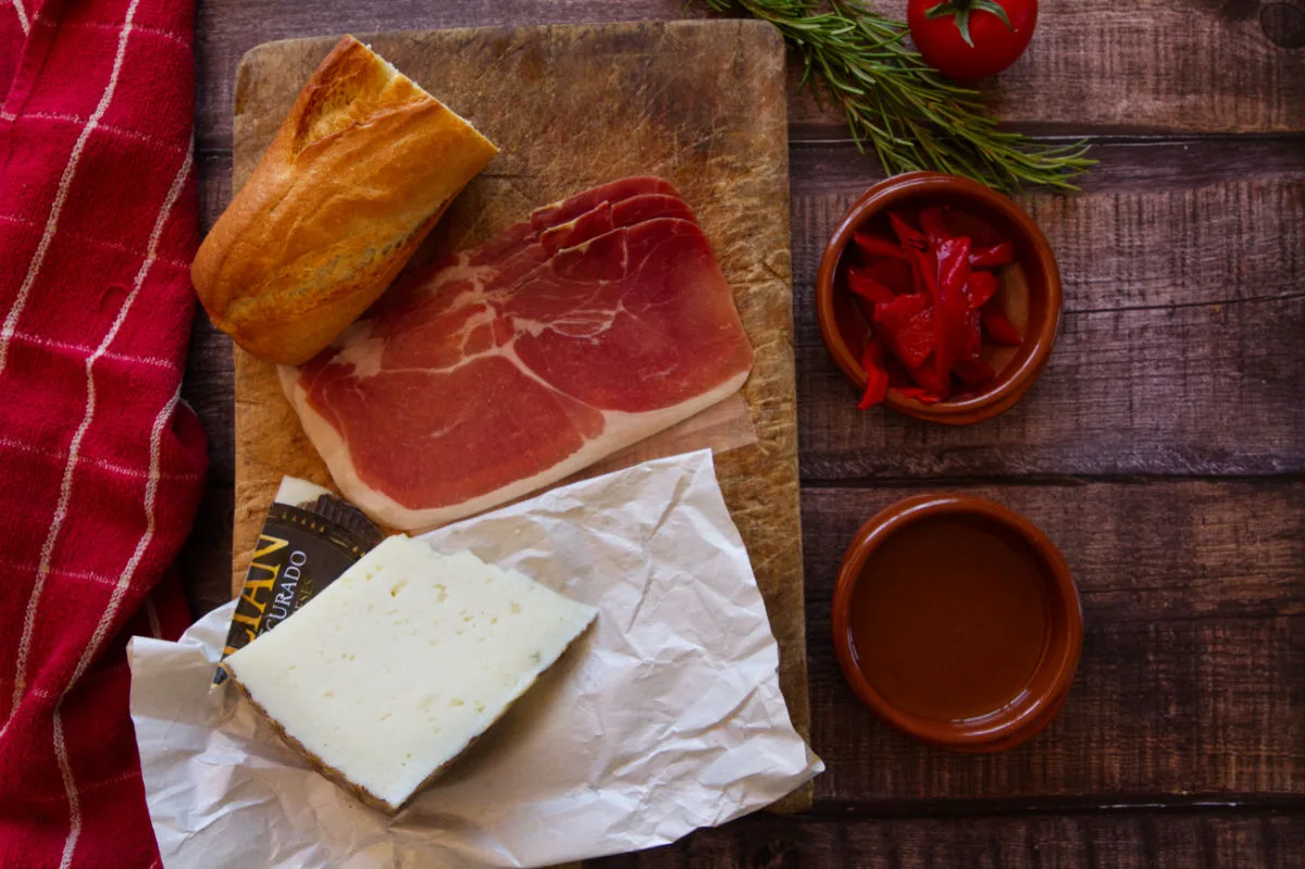 Ingredients for making a ham and mahcego cheese bocadillo are laid out on a kitchen table.