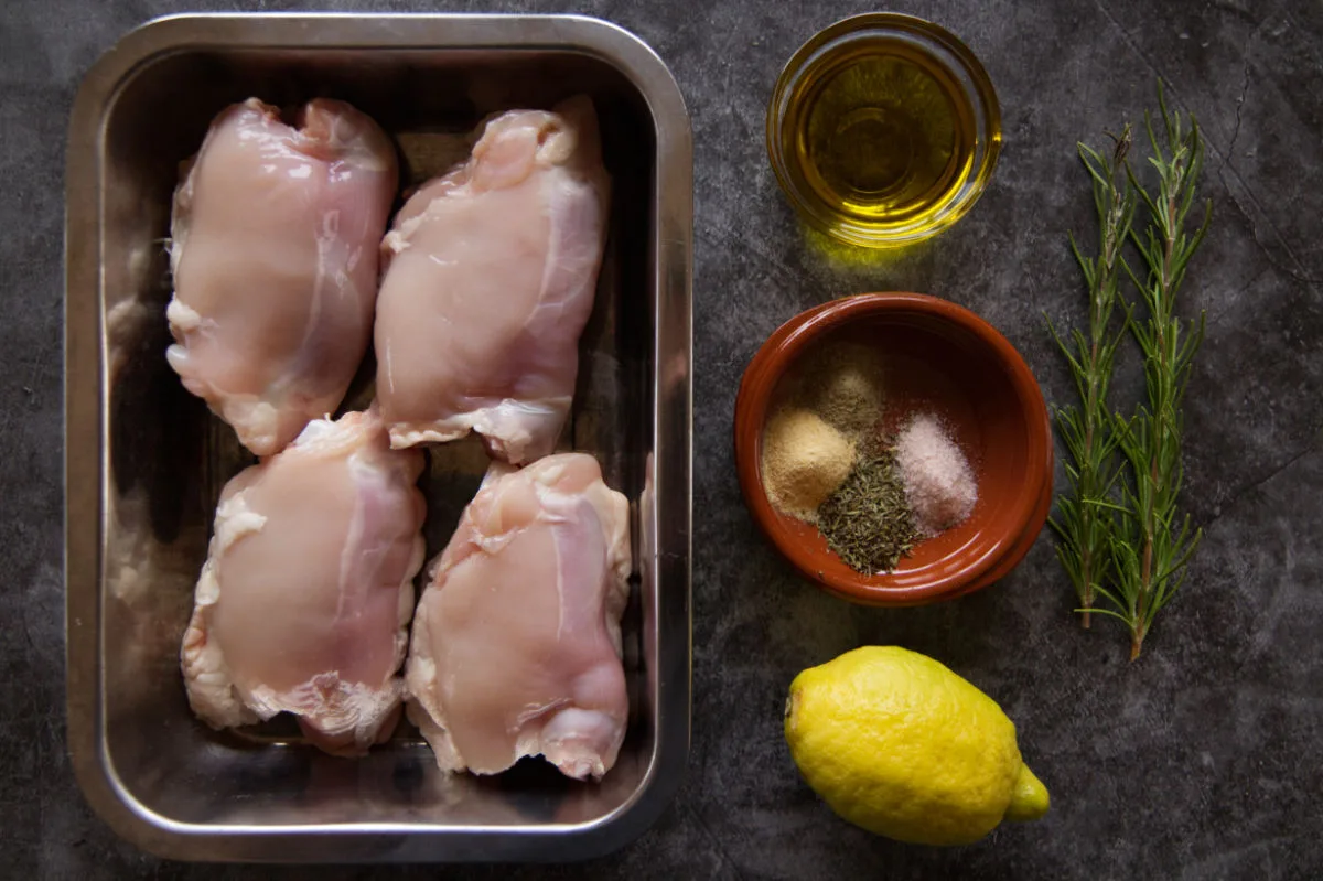 Chicken thighs sit in a baking tray beside a lemon, some spices, and olive oil. 