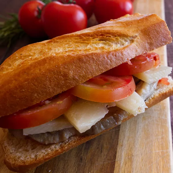 A pork loin bocadillo sits on a chopping board beside some ripe tomatoes