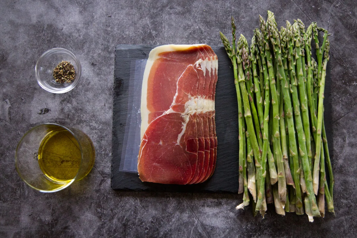 Some sliced Serrano ham sits beside a bunch of asparagus, some olive oil, and some cracked black pepper. 