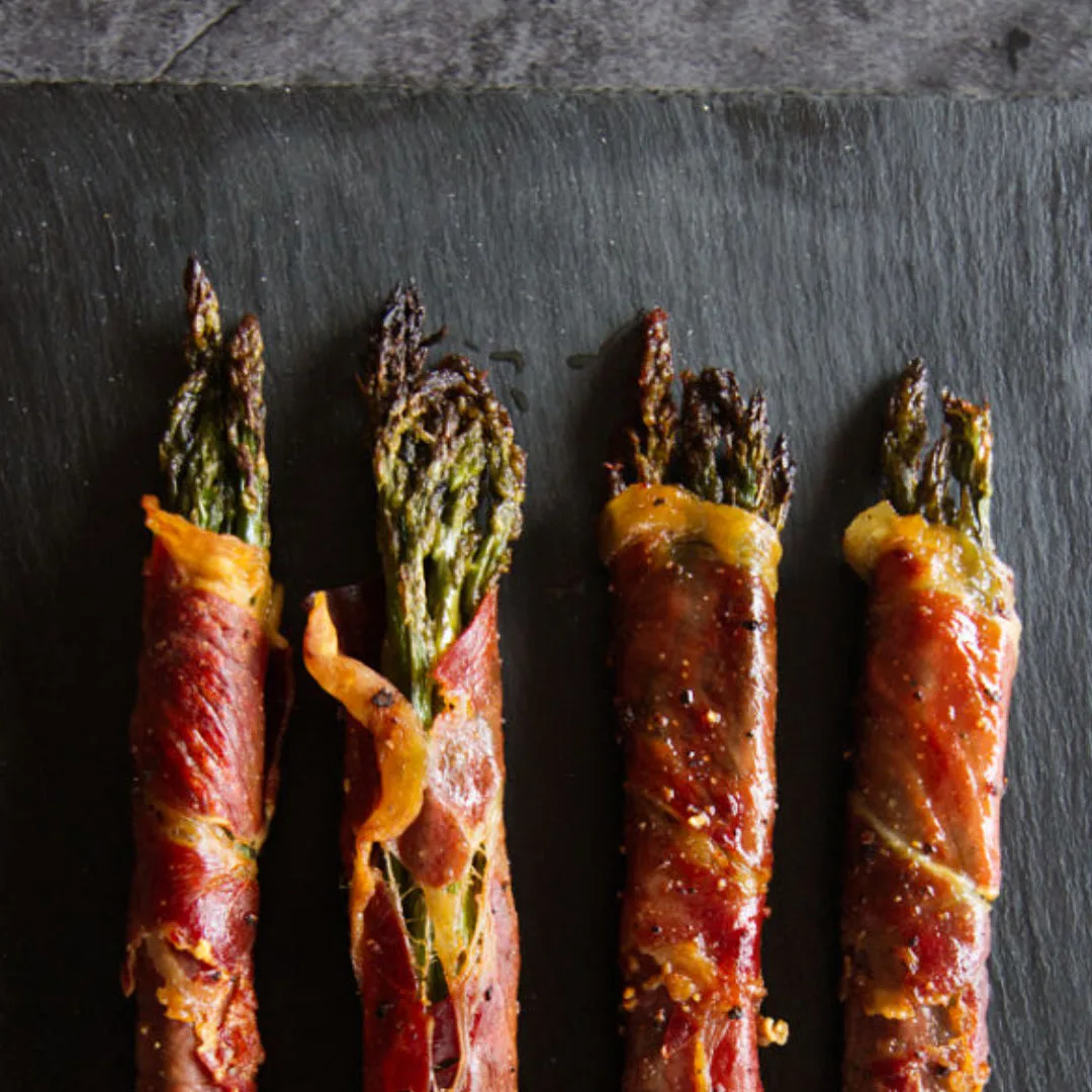 Roasted asparagus spears wrapped in Serrano ham sit on a slate plate.
