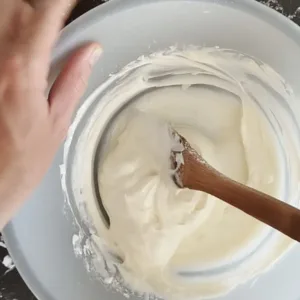 Cream cheese, confectioers sugar, and double cream are mixed by hand in a large white bowl.