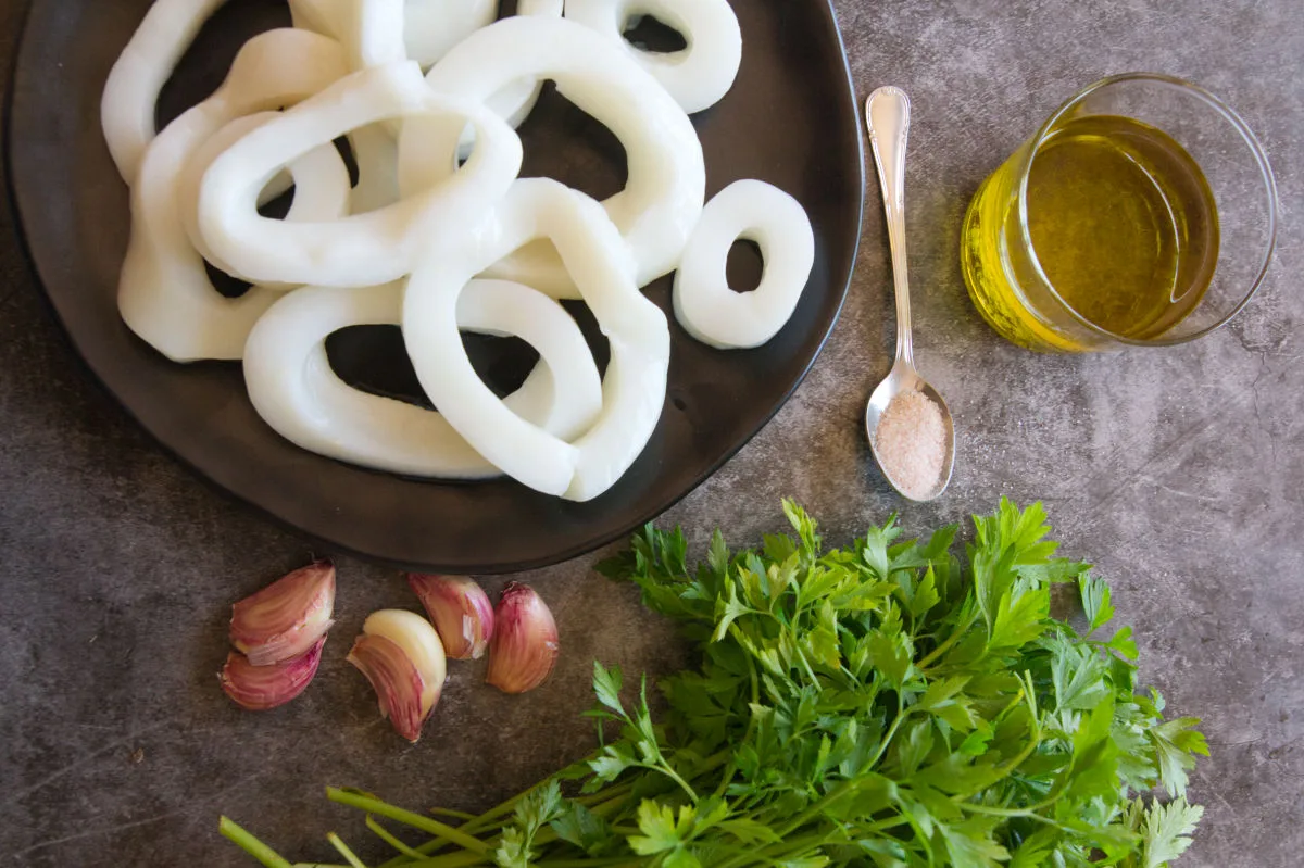Some large squid rings sit beside some garlic, fresh parsley, and olive oil. 