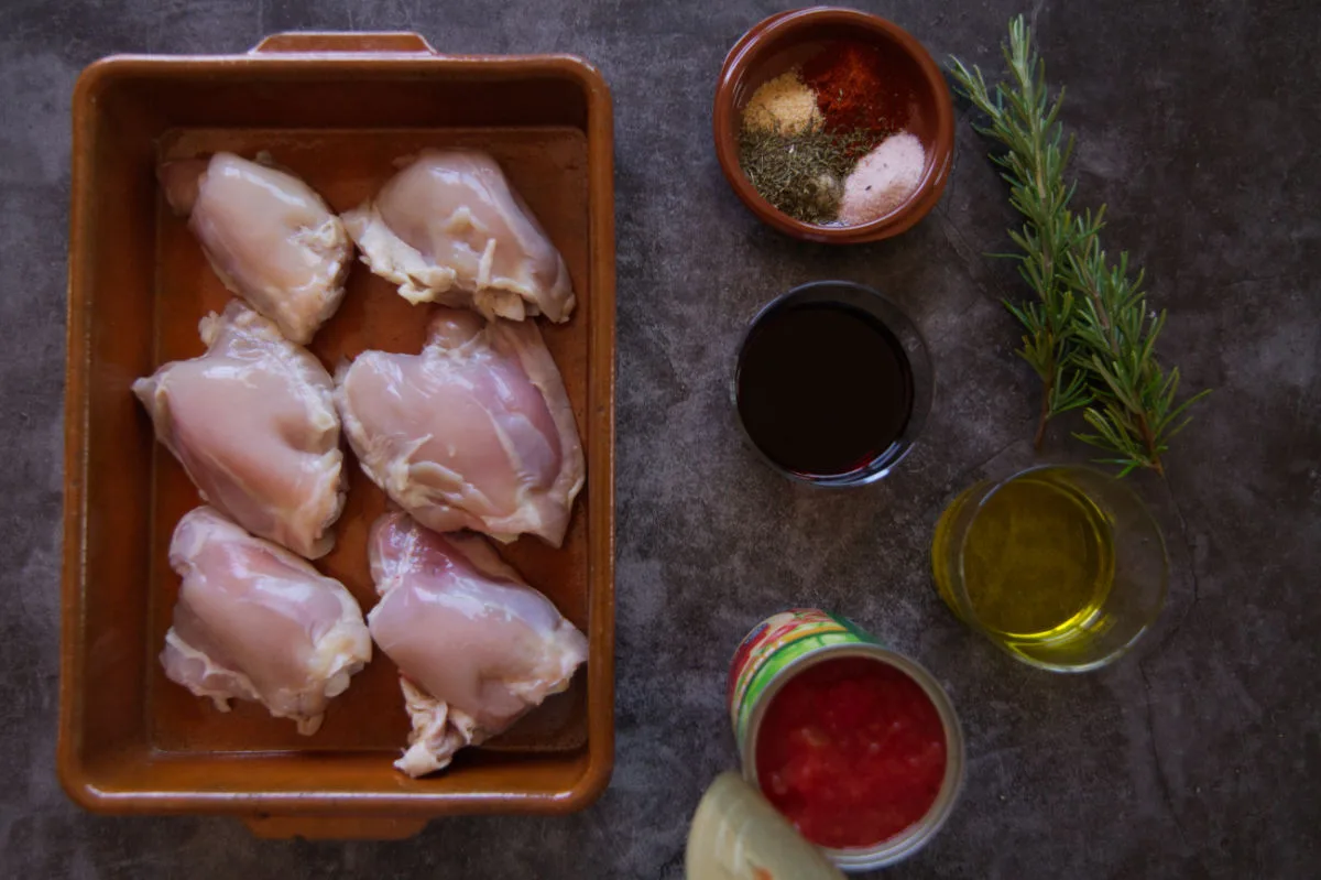 Uncooked chicken thighs sit in a dish beside tinned tmoatoes, herbs, spices, and red wine. 