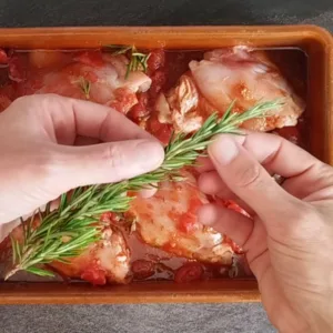 A sprig of rosemary is added to a pan of chicken thighs.