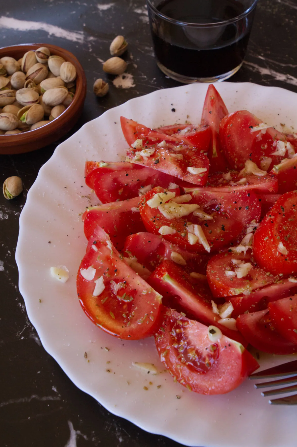 A plate of tomato salad sits beside some pistachio nuts and a glass of red wine.