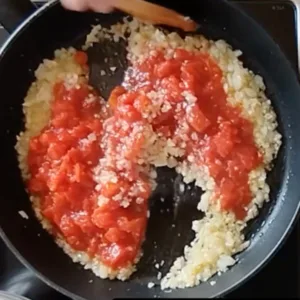 A can of tomatoes is added to a pan of sauteed onions and garlic.
