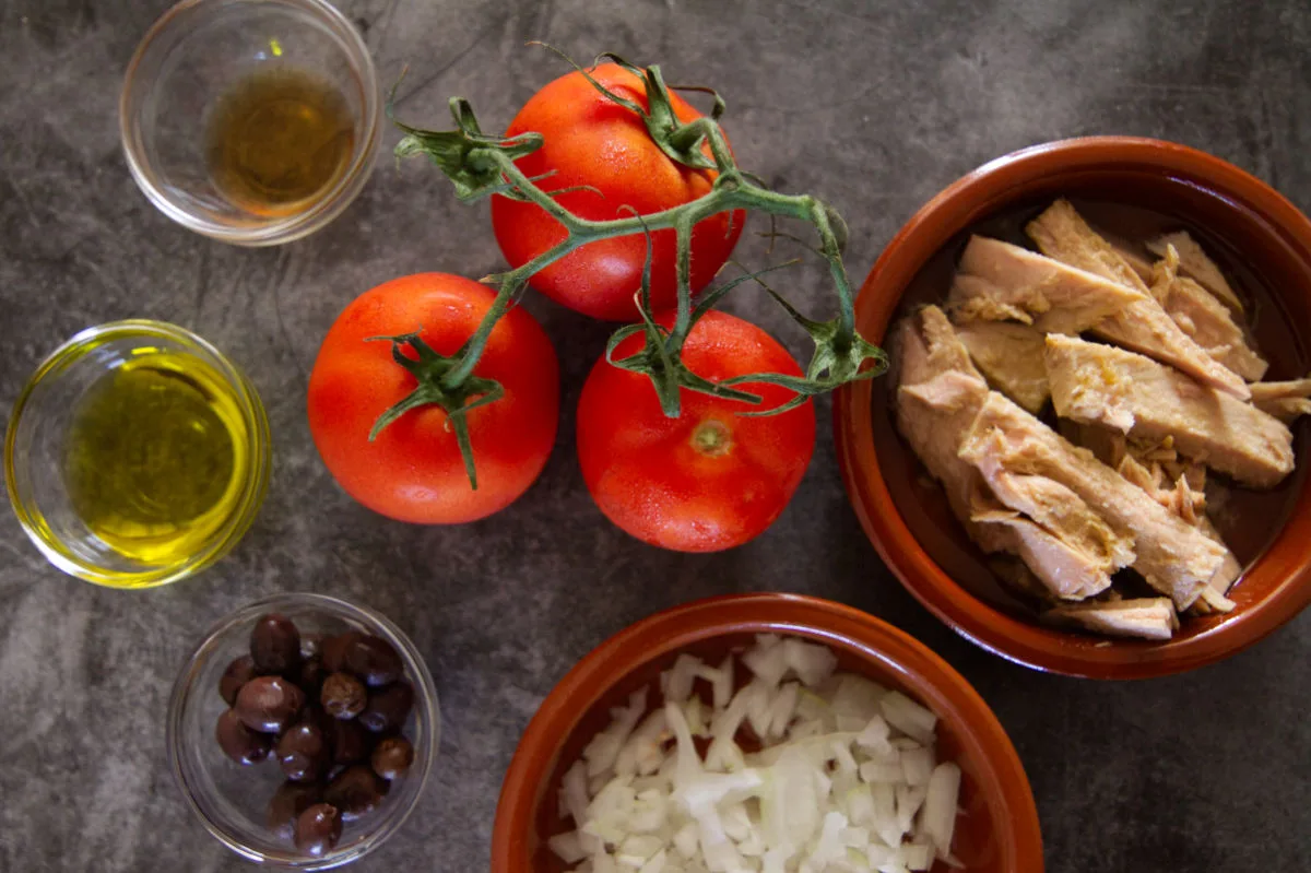A bowl of tunna, some tomatoes, olive oil, olives, and seasoing sit on a counter top. 