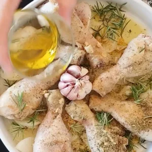 A head of garlic in a dish with some fresh rosemary and chicken drumsticks.