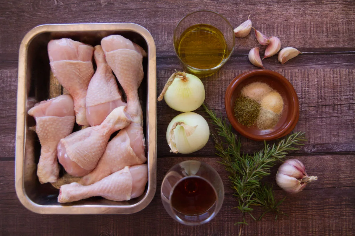 A tray of chicken drumsticks next to some onions, garlic, and fresh rosemary.