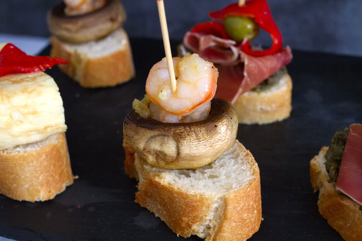 A plate of pintxos with a mushroom and shrimp stacked on a piece of bread.