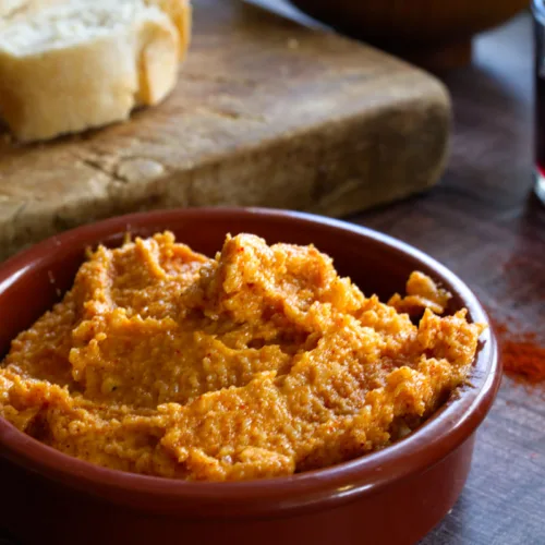 A bowl of Almogrote spiced cheese dip sits beside some slices of bread and some red wine.