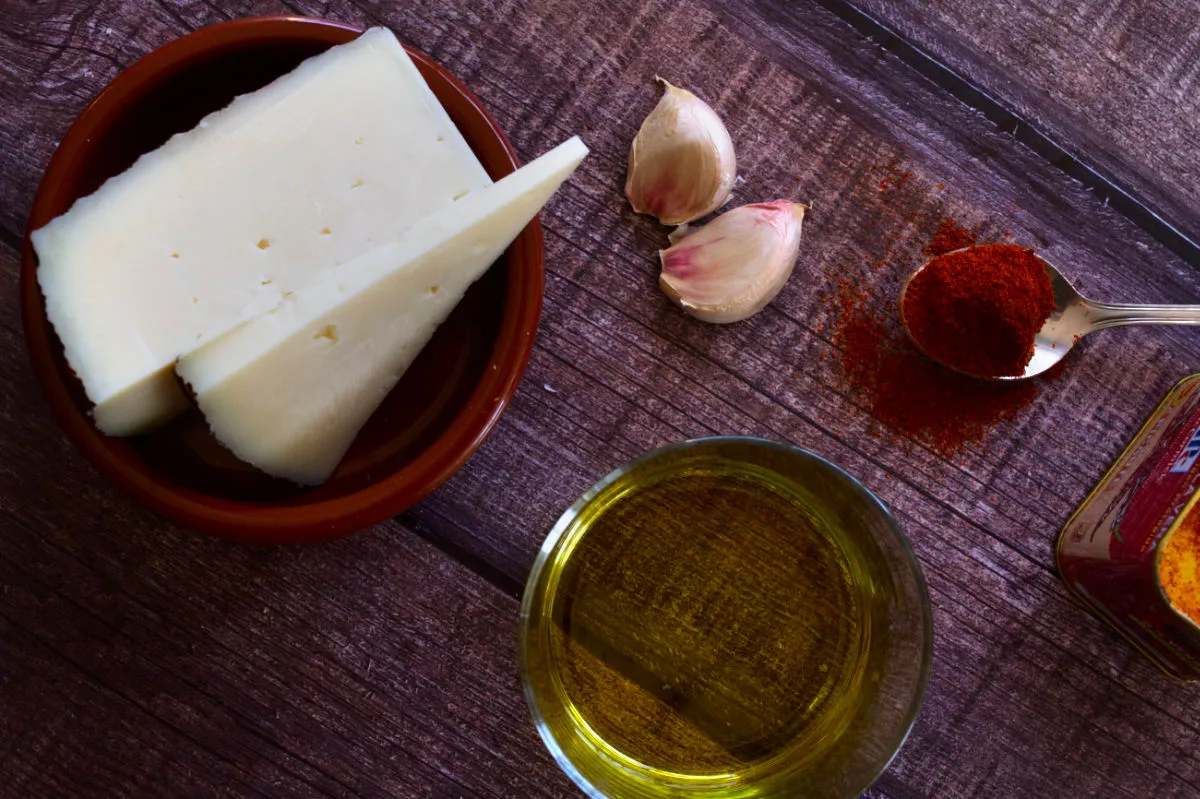A block of cheese is laid next to some olive oil, garlic, and smoked paprika. 