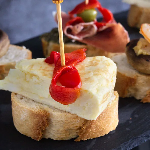 A slice of otrtilla sits on a slice of bread skewered with a ribbon of red pepper on top. 