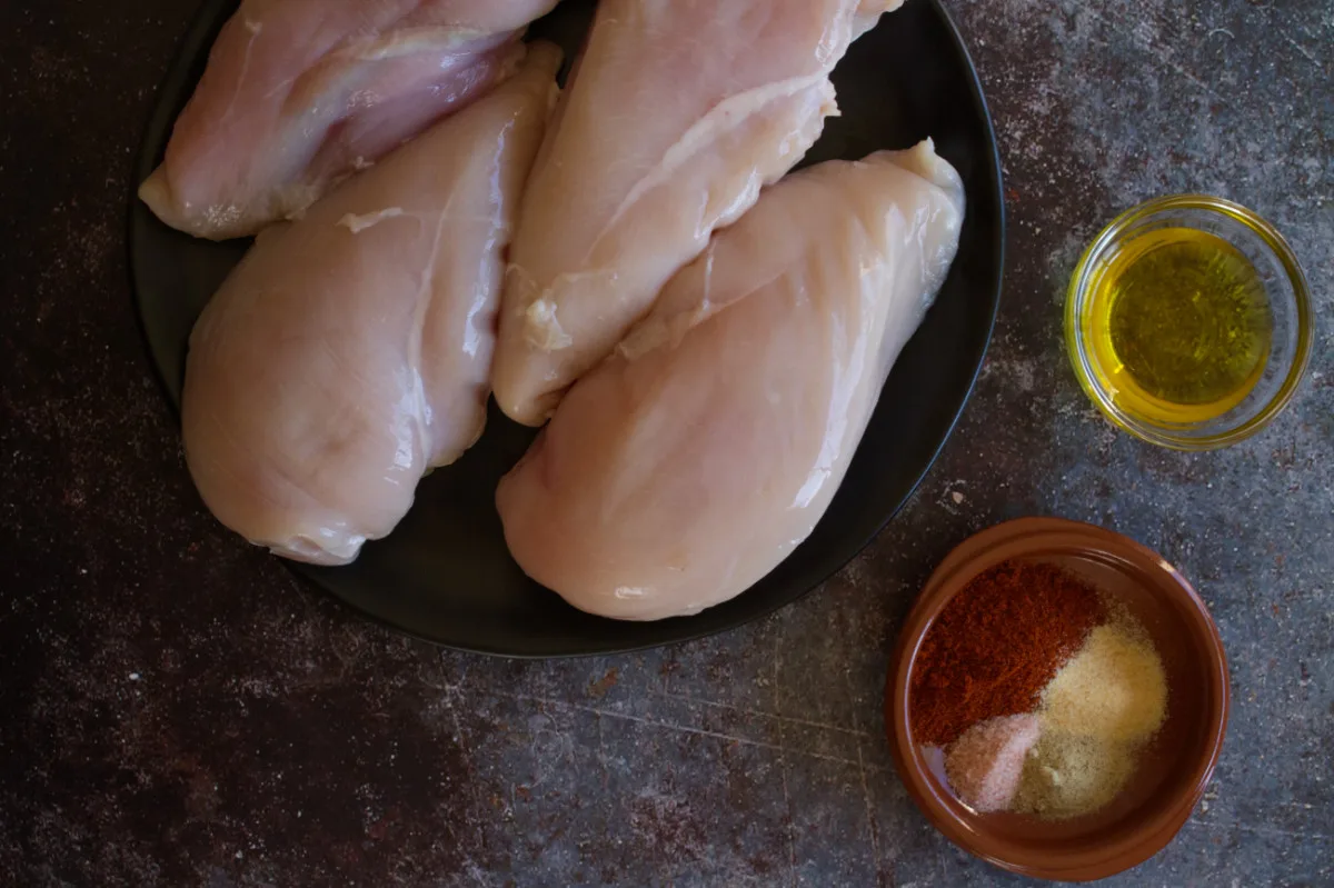 Chicken breasts sit beside some spices and olive oil. 