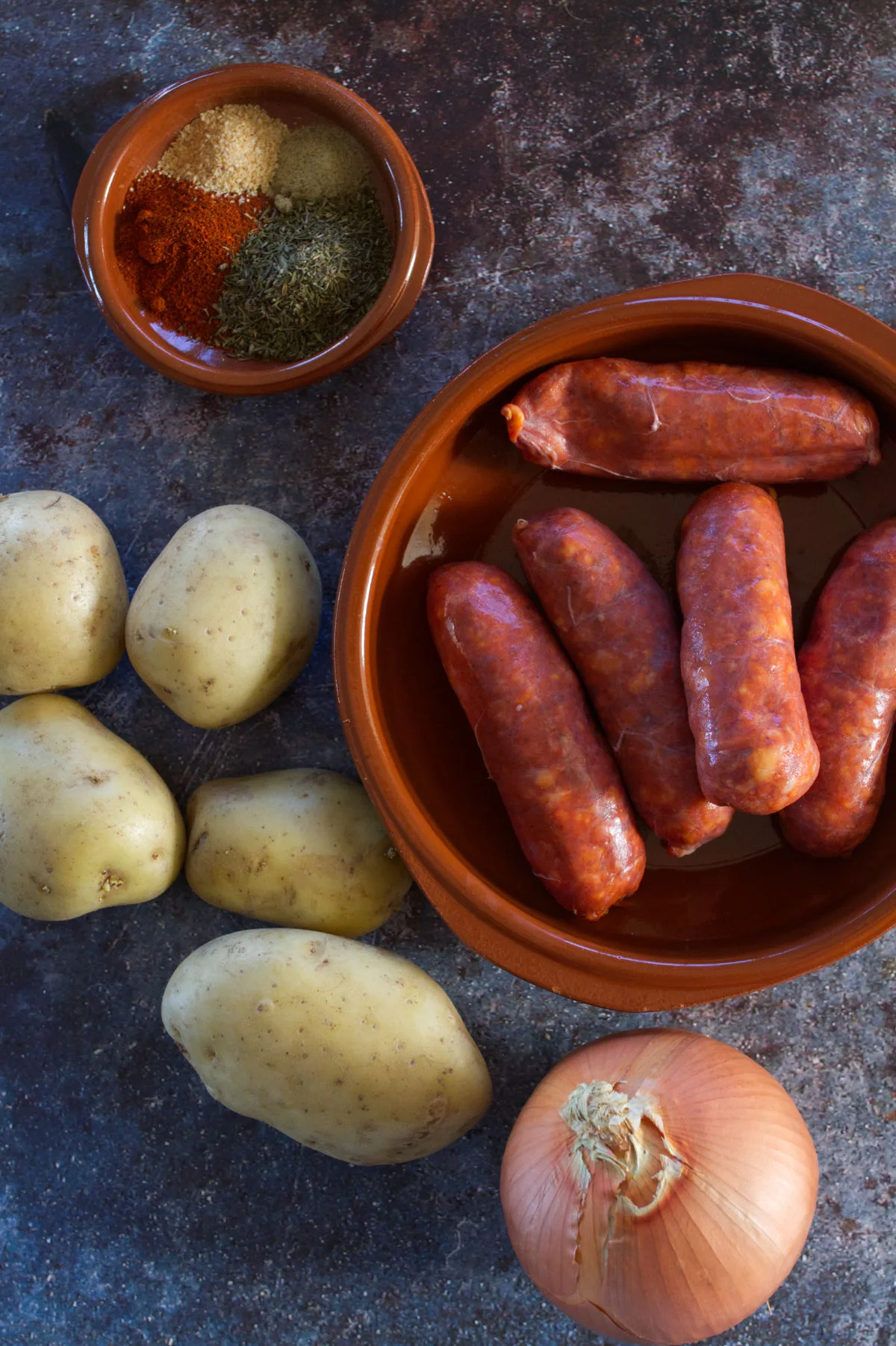 A bowl of chorizo sits next to some potatoes, onion, garlic, and spices. 