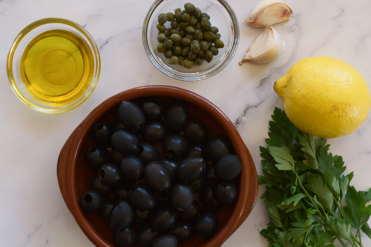 A bowl of olives sits next to some olive oil, capers, lemon, and fresh parsley.