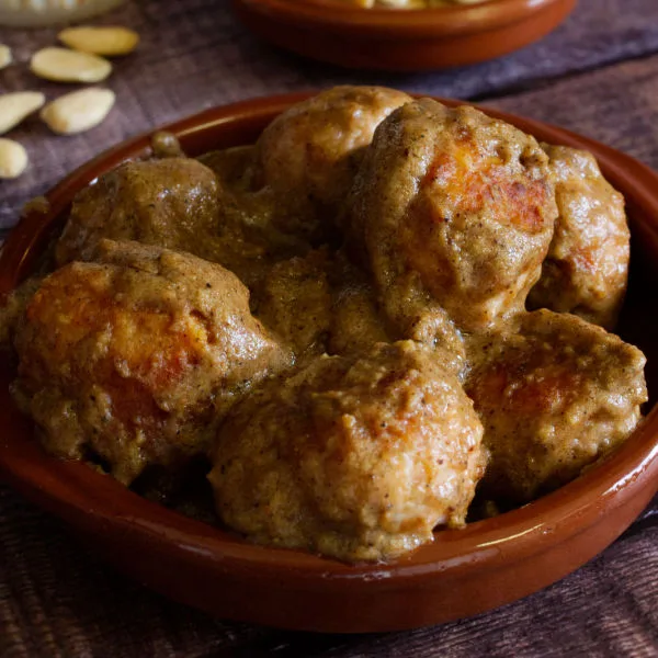 A small tapas serving of Chicken meatballs with almond sauce sits beside some amonds and aglass of red wine.