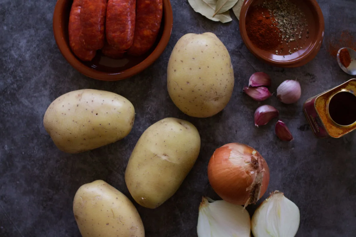 Some large potatoes sit beside chorizo sausages, onions, garlic cloves, and paprika. 