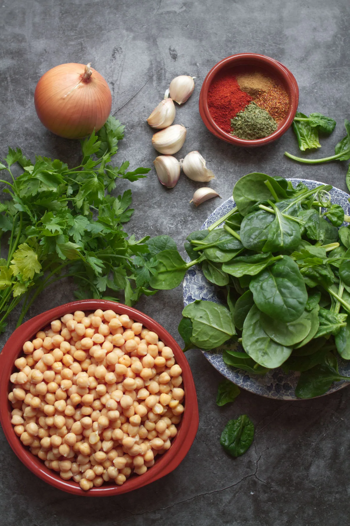 A alrge bowl of chickpeas sists beside a bowl of spinach, onion, garlic, and some herbs and spices. 