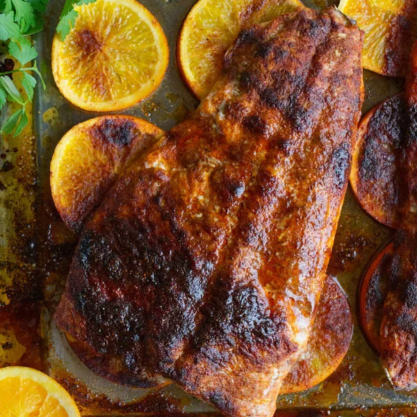 A large fillet of Spanish-style spicy salmon beside some orange rounds and fresh herbs.