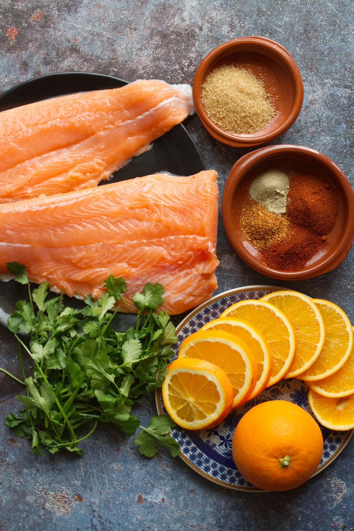 large raw salmon fillets sit beside some orange slices, spices, and fresh herbs. 