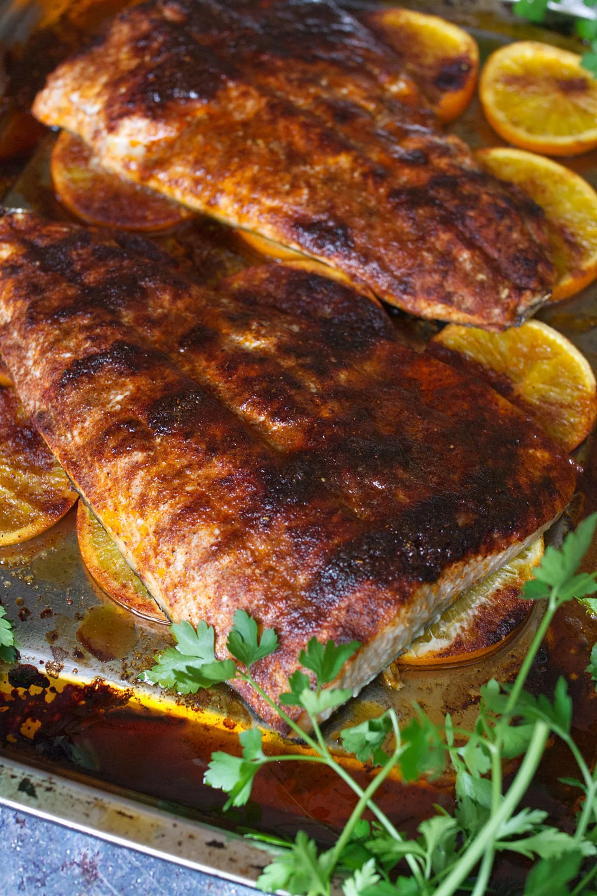 Large fillets of Spanish-style spicy salmon beside some orange rounds and fresh herbs.