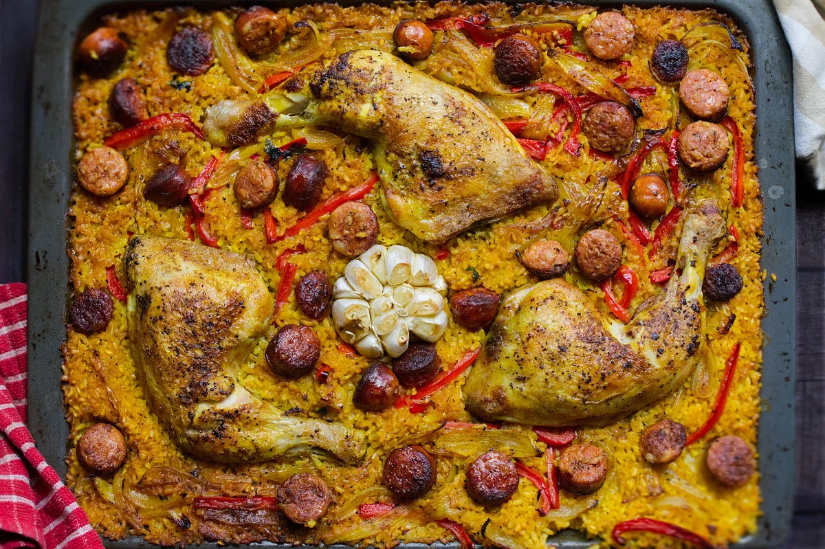 A large sheet pan with chicken chorizo and rice, and garnished with a whole head of garlic.
