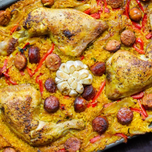 A large sheet pan with chicken chorizo and rice, and garnished with a whole head of garlic.
