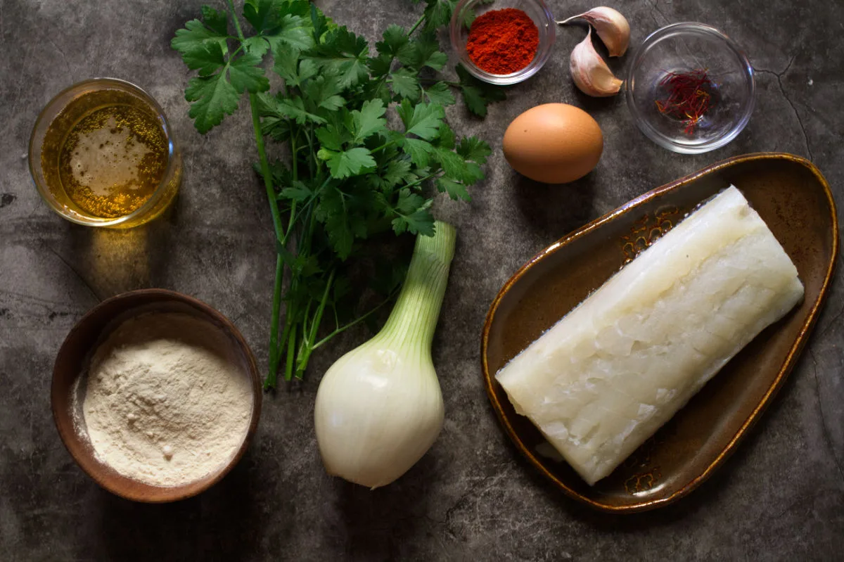 A large fillet of cod fish sits beside some onion, flour, egg, spices, herbs, and beer.