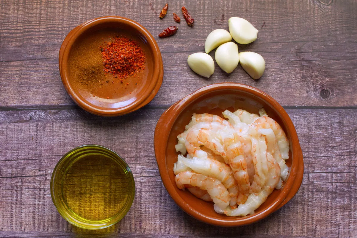 A plate of large prawns sits beside some spices, olive oil, garlic, and chili.