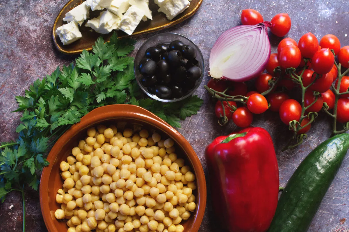 a large bowl of chickpeas sits beside red bell pepper, cucumber, cherry tomatoes, feta cheese, and herbs. 